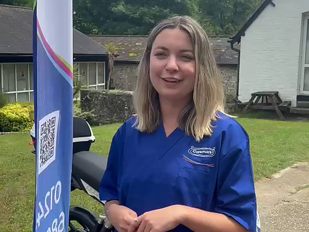 A Caremark care assistant in uniform smiling beside an Eskuta SX-250, with a Caremark banner alongside, outside a Caremark Arun site