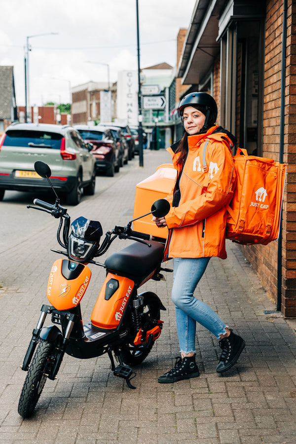 Person in orange delivery jacket with backpack standing next to an electric scooter on a city street.