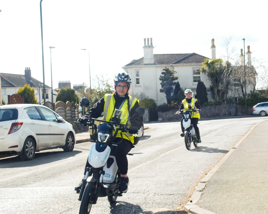 Two carers in hi-vis riding liveried Eskuta SX-250s along a residential street