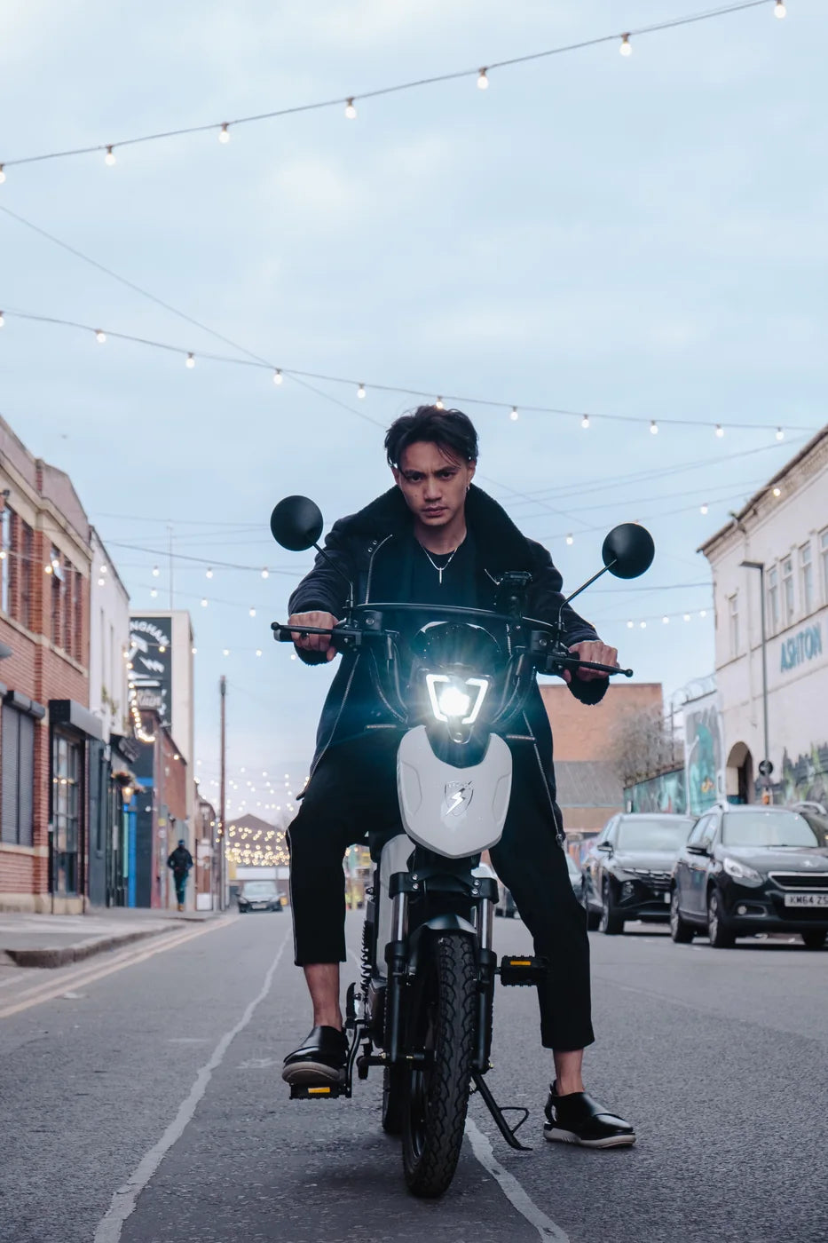 A rider on a white Eskuta SX-250 head-on in a city street at dusk, headlight lit, mirrors, forks and pedals visible