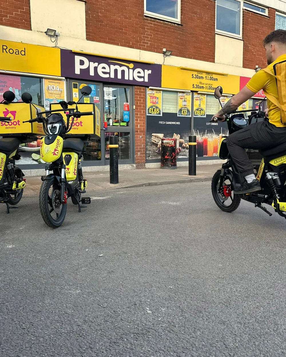 A delivery rider setting off from a convenience store on an Eskuta SX-250 with an insulated delivery bag, two more bikes waiting outside